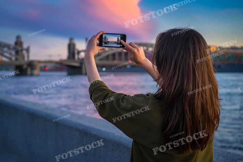 На этом фото девушка фотографирует городской пейзаж на свой смартфон. Она стоит спиной к камере, на фоне закатного неба с яркими красками и реки, видны очертания моста. Из-за наличия воды и неба, фотография выглядит мирно и расслабляюще, и может быть использована в статьях о путешествиях, в блогах о лайфстайле, или как иллюстрация к темам о фотографии и технологиях.